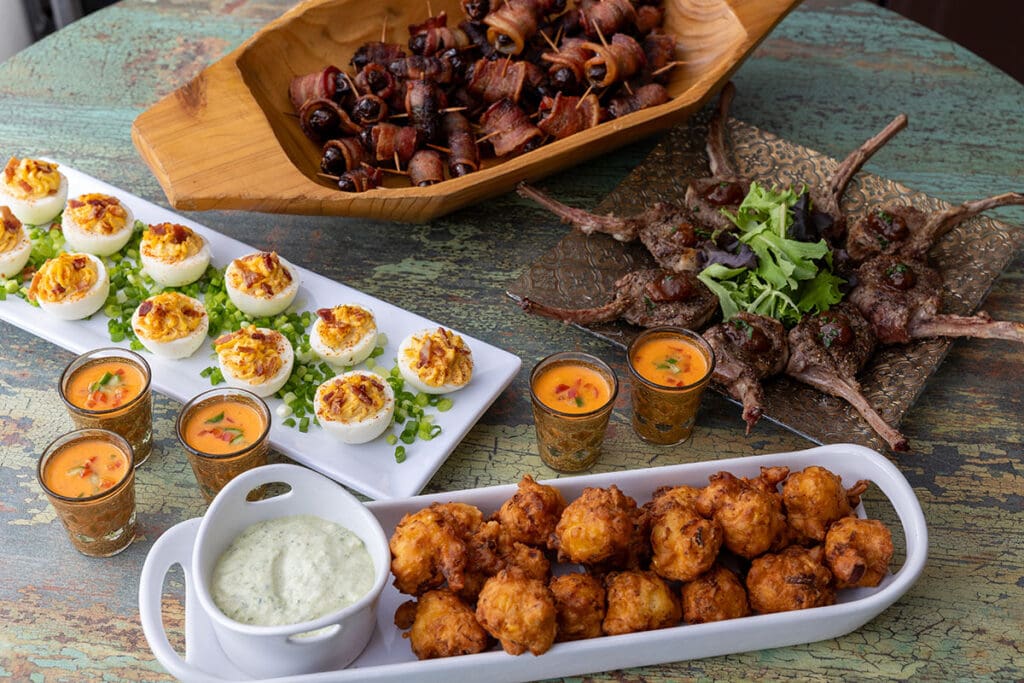 Overhead view of several food items in serving dishes: Bacon-wrapped dates, deviled eggs, lamp chops, gougeres, and soup
