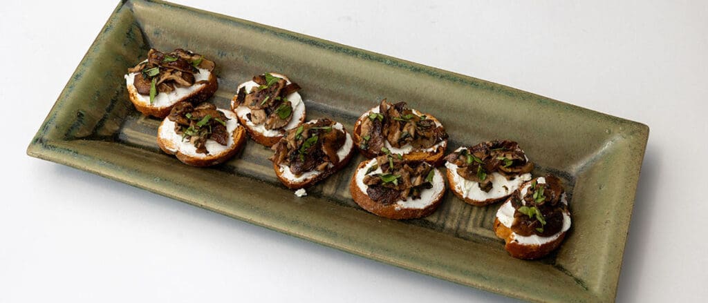 Small toasted breads with spread and mushrooms on them, with herbs on top on a long rectangle platter