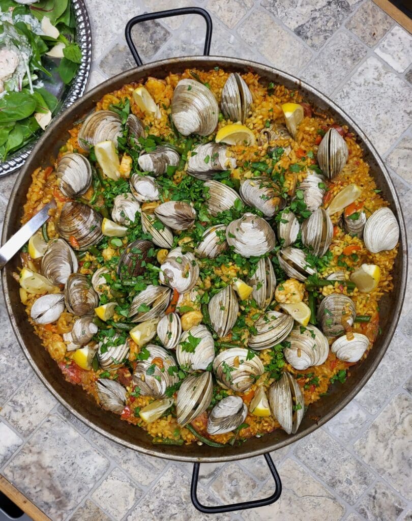 A paella platter with paella that includes clams, lemon wedges and cilantro garnish