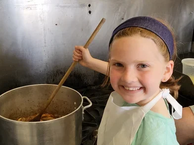 Child smiling at the camera, while they stir a pot of soup with a wooden ladle
