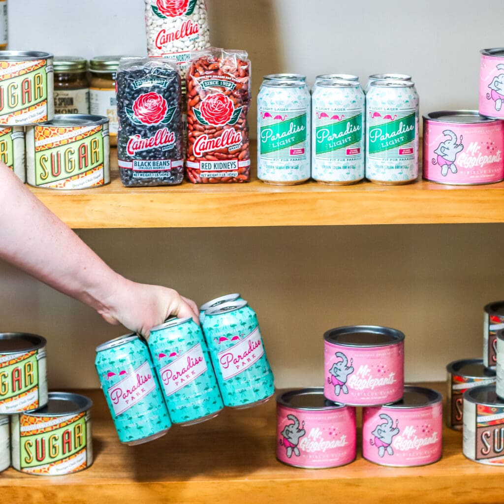A person picks up a six-pack of beer cans from the colorful, well-stocked shelves at Nolavore.